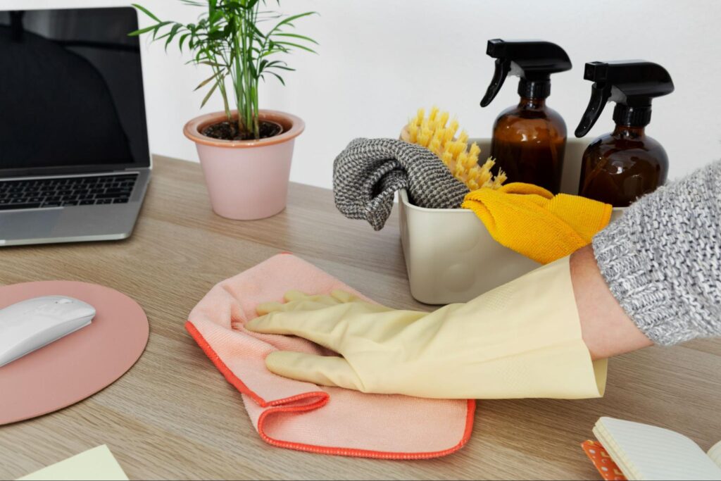 Gloved hand wiping an office desk beside cleaning supplies.